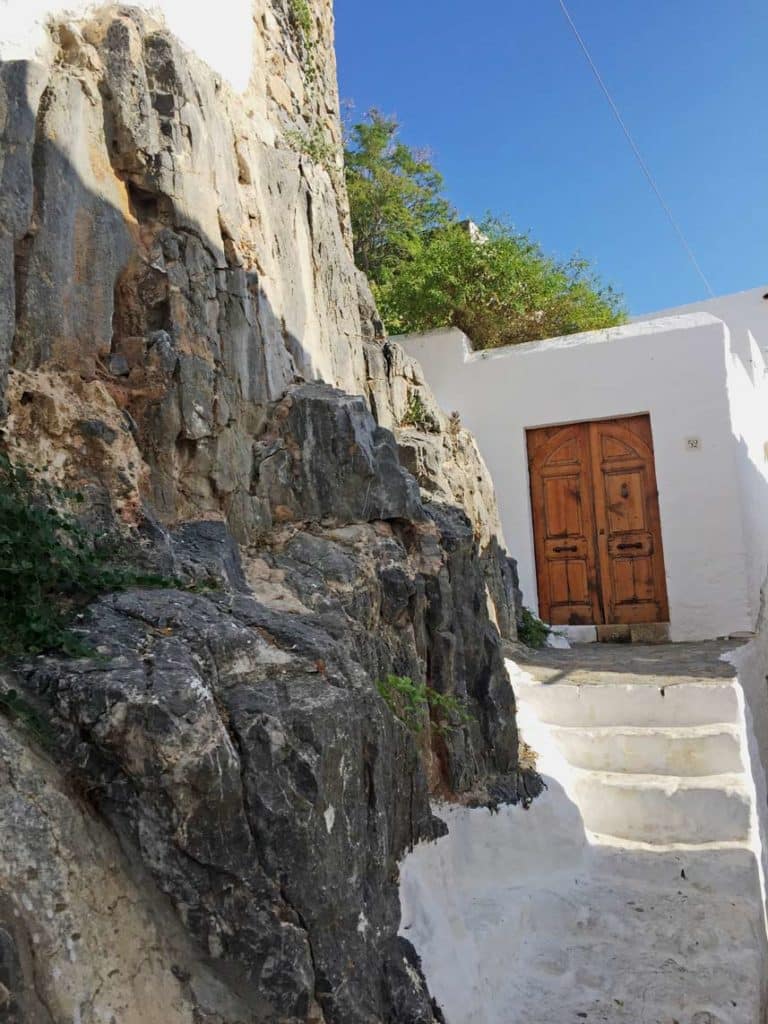 weisses-gebaeude-mit-brauner-tuer-an-treppe-und-felsklippe-in-lindos-auf-rhodos