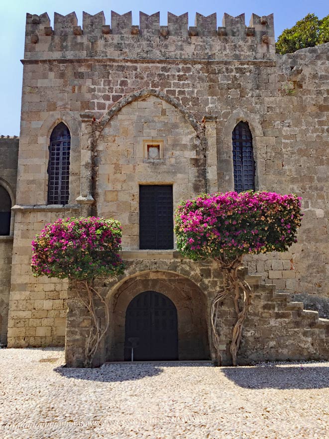 steinernes-gebaeude-auf-rhodos-mit-zwei-lila-bluehenden-baeumen-davor