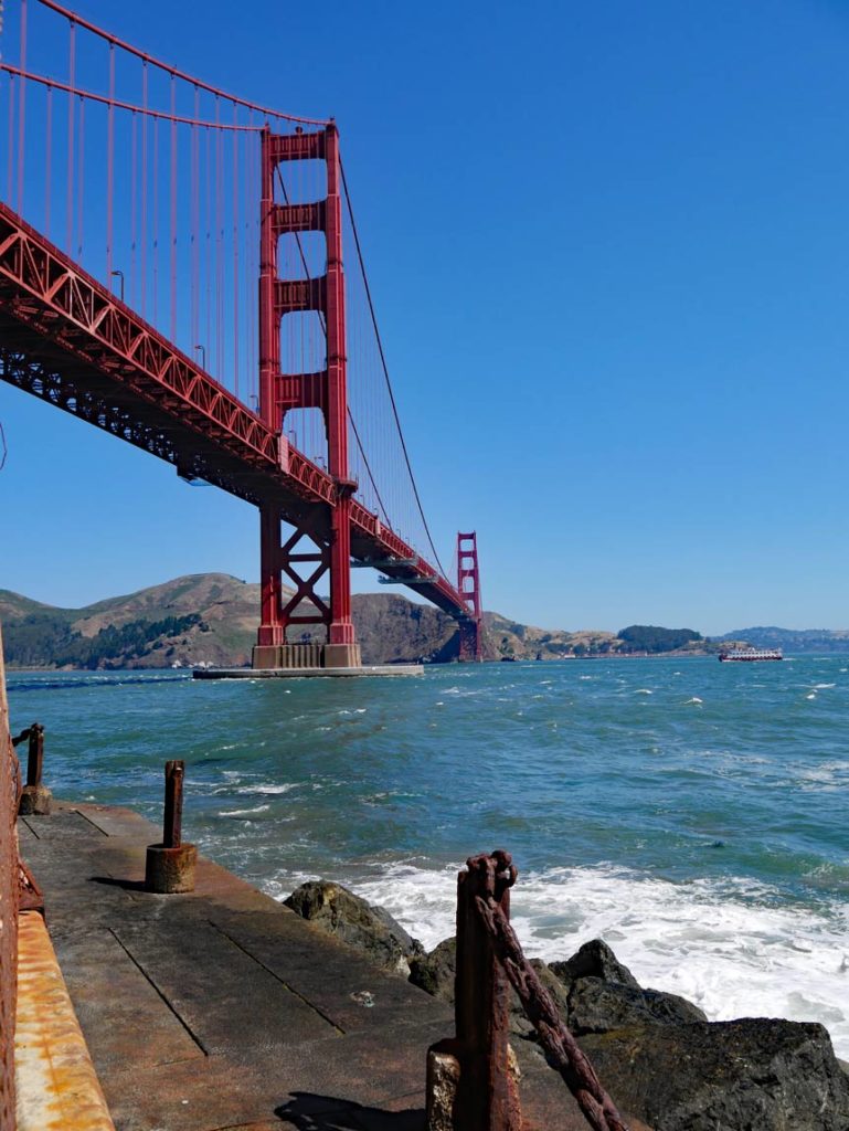 rostige-metallabsperrung-vor-dem-wasser-der-fotografierende-steht-fast-direkt-unter-golden-gate-bridge-die-in-den-huegeln-sausalitos-endet