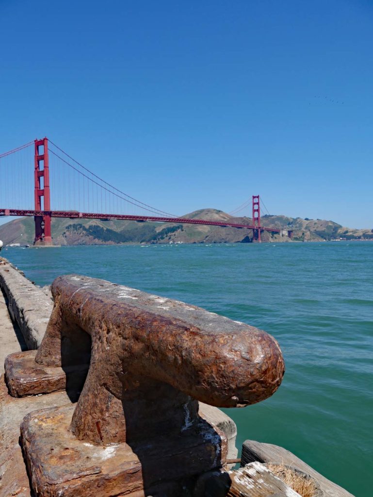 vorne-im-bild-ein-verrosteter-poller-dahinter-blaues-wasser-und-schliesslich-die-rote-golden-gate-bridge