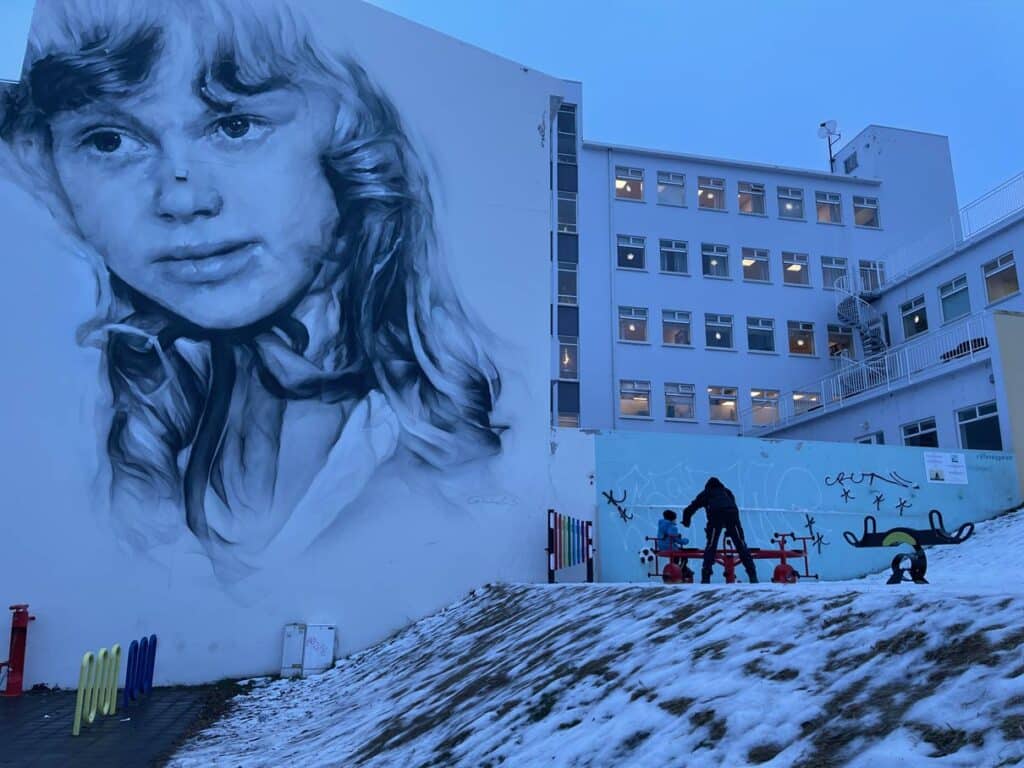 16 Nordisland Sehenswürdigkeiten, die ihr nicht verpassen dürft! kinder-spielen-auf-spielplatz-im-akureyri-vor-gebaeude-mit-grossem-streetartbild-eines-maedchengesichts-in-der-daemmerung