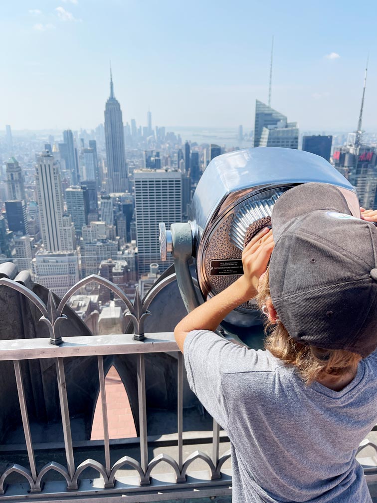 kind-blickt-durch-fernglas-vom-rockefeller-center-top-of-the-rock-aussichtsplattform-auf-das-empire-state-building-new-york-city-hochhaeuser-skyline