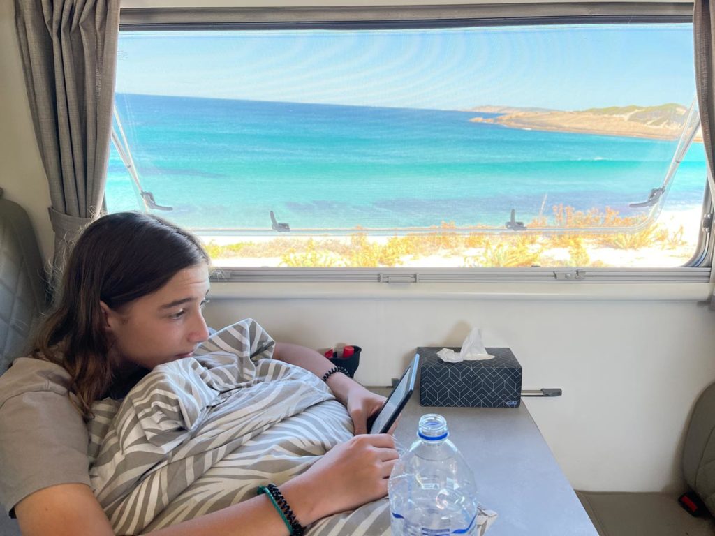 teenager-liest-auf-ebook-im-wohnmobil-vor-dem-fenster-traumstrand-in-australien