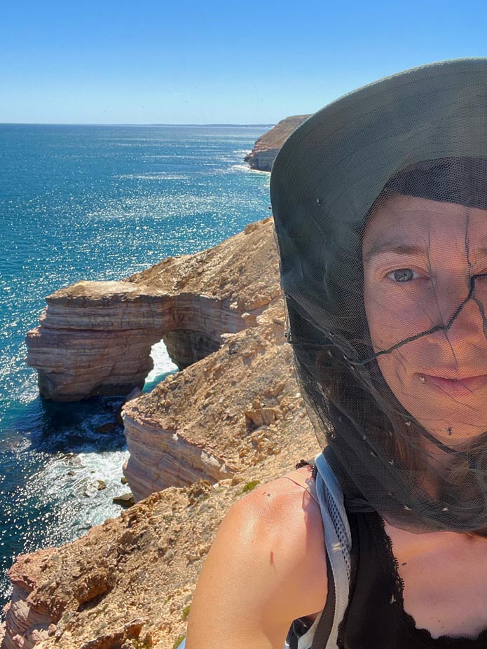 Ultimativer Guide für den Westaustralien Roadtrip frau-mit-moskitohut-an-felsenkueste-mit-felsentor-im-kalbarri-nationalpark-rundherum-viele-buschfliegen