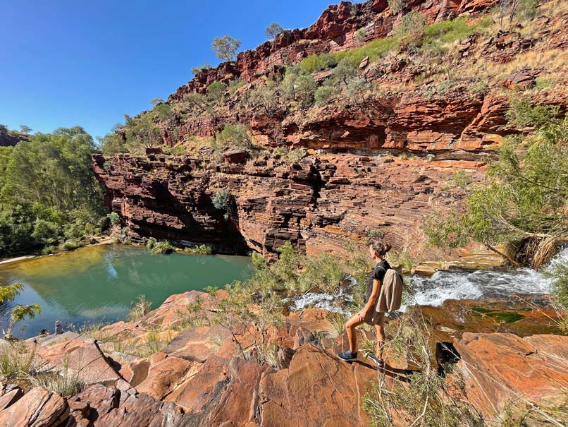 frau-steht-oben-an-fortescue-falls-karjini-nationalpark-rote-sandsteinlandschaft-mit-gruenem-see-am-wasserfall