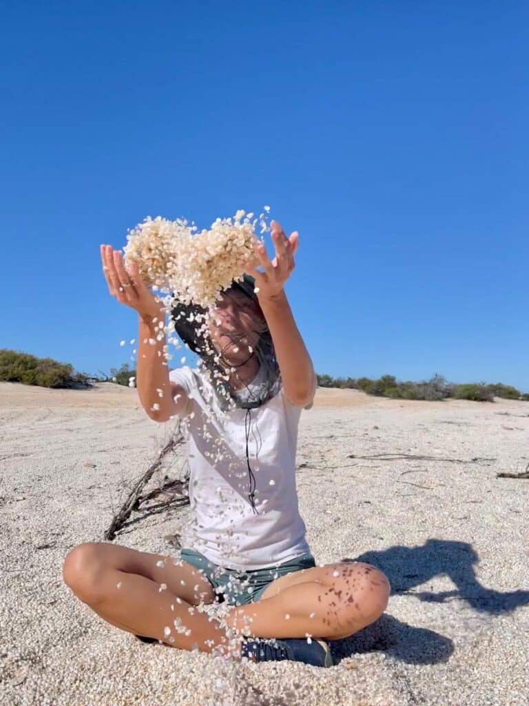 jenni-von-der-strandfamilie-sitzt-auf-muschelstrand-im-schneidersitz-und-wirft-weisse-muscheln-in-die-luft-welche-ein-herz-formen