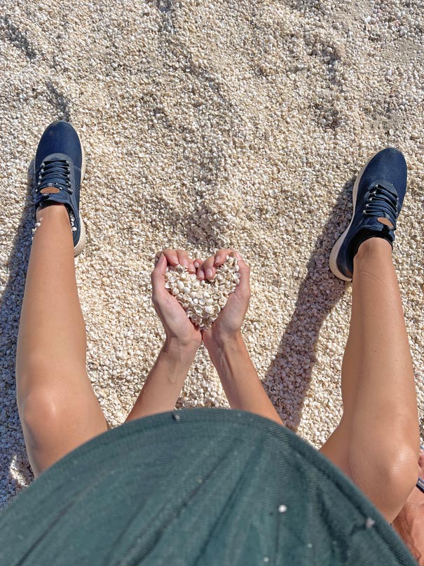 fotografie-von-oben-auf-frau-die-auf-weissem-muschelstrand-sitzt-man-sieht-nut-ihren-hut-die-beine-und-in-den-haenden-haelt-sie-muscheln-zu-herz-geformt