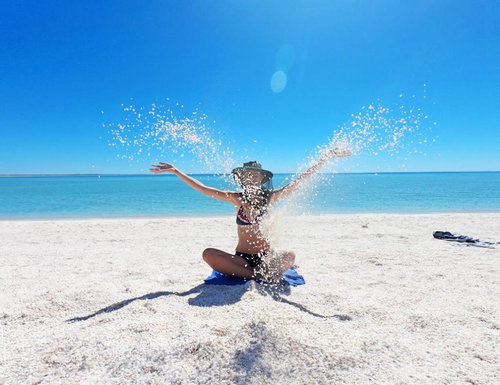 frau-sitzt-auf-weissem-muschelstrand-im-bikini-am-shell-beach-in-westaustralien-und-wirft-muscheln-in-die-hoehe-im-hintergrund-blauer-himmel-und-blaues-meer