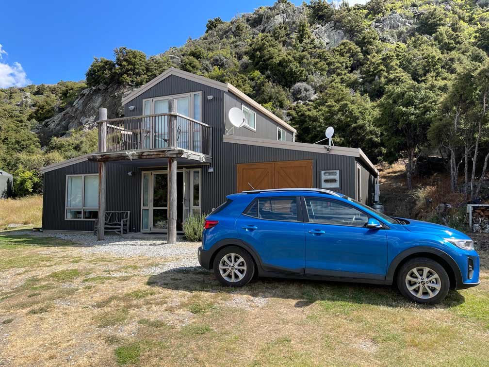 blauer-mietwagen-steht-vor-grauem-ferienhaus-auf-der-wiese-in-neuseeland