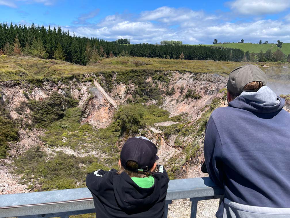 kind-und-vater-blicken-in-rauchende-vulkanische-krater-die-grün-bewachsen-sind-craters-of-the-moon-neuseeland