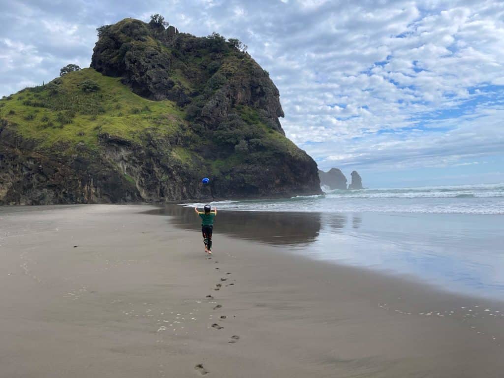 kind-spielt-mit-rugby-auf-schwarzem-strand-vor-felsen-piha-beach-neuseeland-nordinsel-reiseroute
