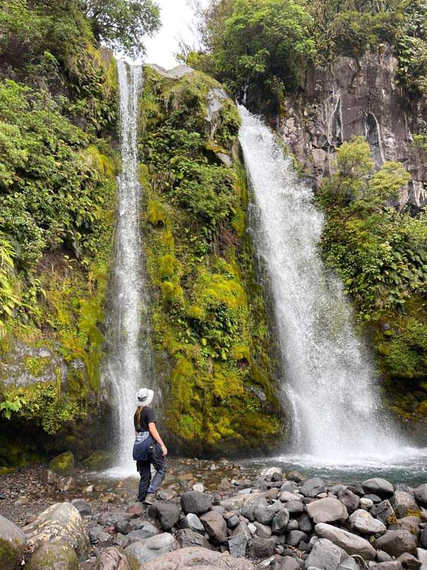 kind-steht-unten-an-einem-zwei-reihigen-wasserfall-in-einer-gruenen-moosigen-landschaft-auf-steinen