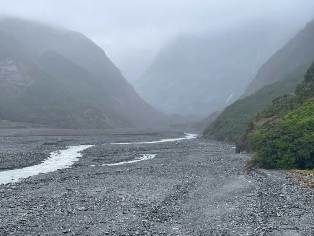 nebel-verdeckt-den-franz-josef-gletscher-sichtbar-ist-nur-die-nahe-umgebung-mit-fluss-im-gestein