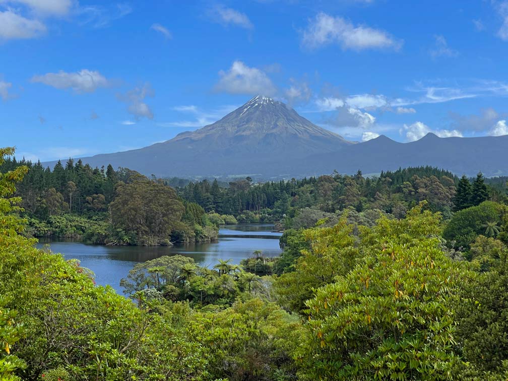 blick-ueber-den-blauen-see-lake-mangamahoe-der-an-den-seiten-dicht-gruen-bewachsen-ist-mittig-der-vulkan-taranaki