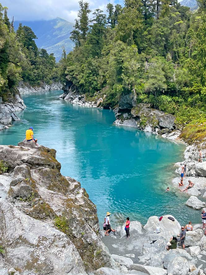 hellblauer-fluss-zwischen-weissem-kalkgestein-dahinter-gruener-wald-touristen-sitzen-am-ufer-der-hokitika-gorge-auf-der-suedinsel-neuseeland