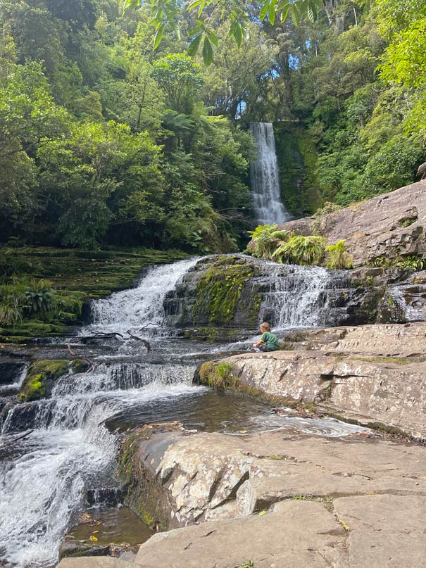 kind-sitzt-neben-mc-lean-falls-der-sich-ueber-mehrere-kaskaden-startend-im-gruenen-wald-dann-ueber-die-felsen-ergiesst-neuseeland