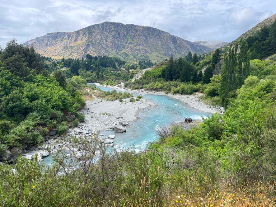 gruene-landschaft-am-rand-im-hintergrund-hoher-berg-mittig-ein-hellblauer-fluss-mit-weissen-steinen-gerahmt-auf-neuseeland-suedinsel