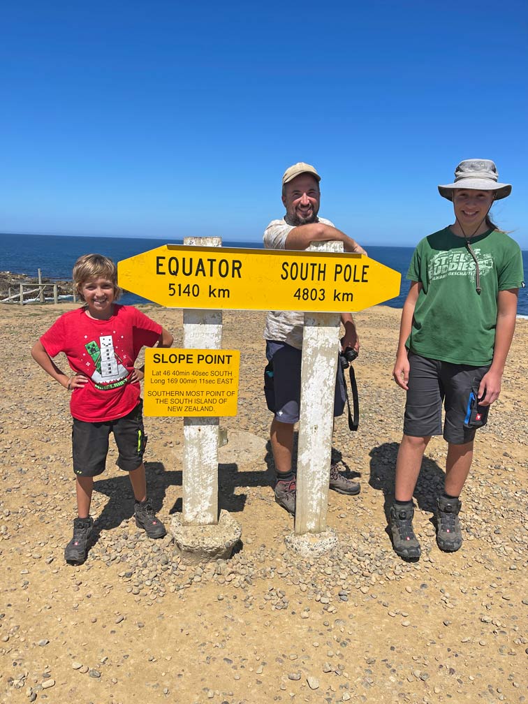 familie-steht-am-gelben-schild-slope-point-mit-richtungsangaben-in-den-catlins-neuseeland