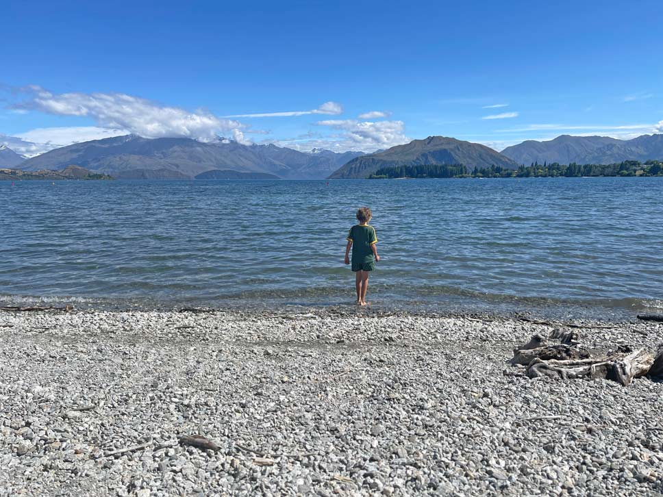 kind-steht-am-steinigen-ufer-am-lake-wanaka-mit-blick-auf-die-berge