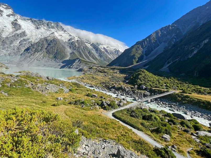 malerische-landschaft-im-mount-cook-nationalpark-gruene-landschaft-mit-haengebruecke-ueber-den-fluss-und-schneebedeckten-bergen