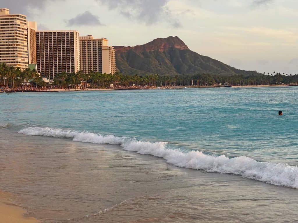 wellen-am-sandstrand-und-hellblaues-meerwasser-am-waikiki-beach-im-hintergrund-hochhaeuser-und-bergekette-diamond-head