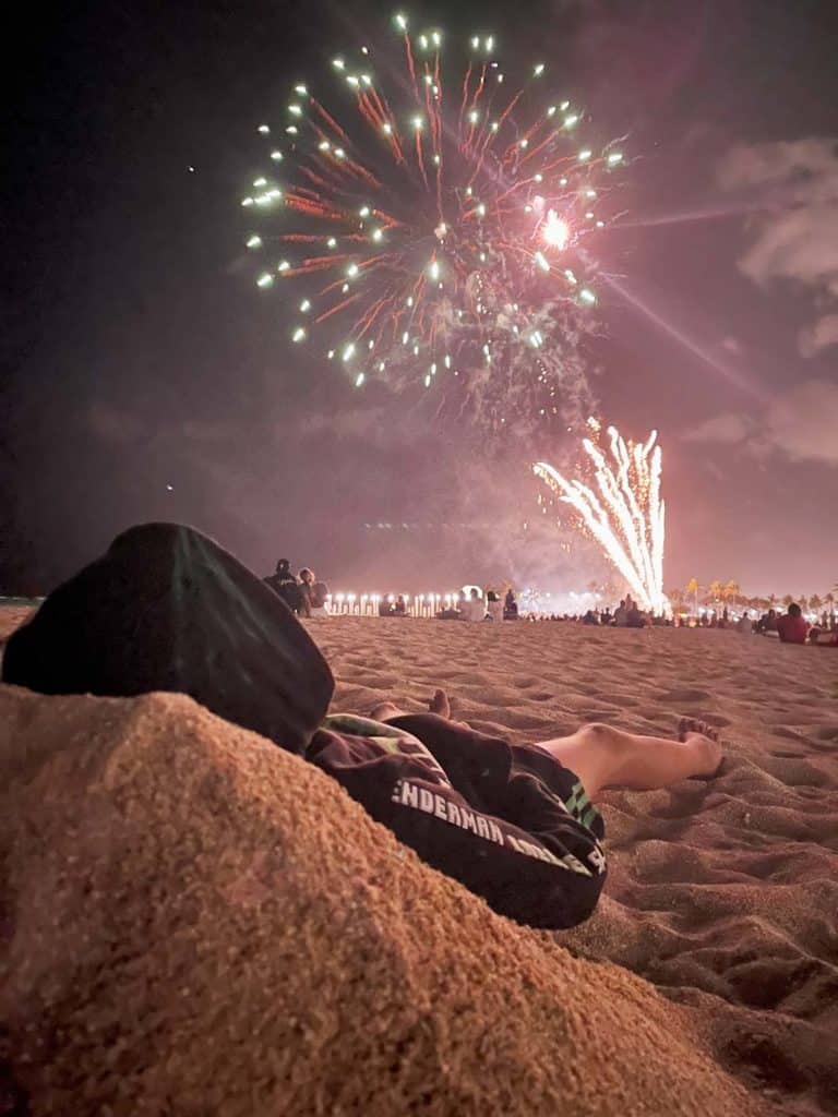 kind-liegt-im-sand-und-schaut-feuerwerk-am-himmel-am-waikiki-beach