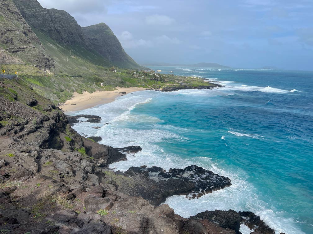 aussicht-ueber-schwarze-lava-die-sich-ins-meer-ergiesst-hellen-sandstrand-und-gruene-berge-auf-oahu