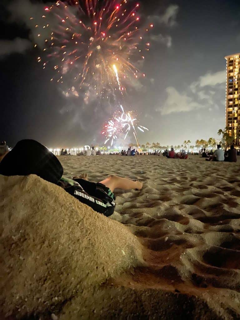 kind-liegt-im-sand-am-waikiki-beach-und-beobachtet-feuerwerk