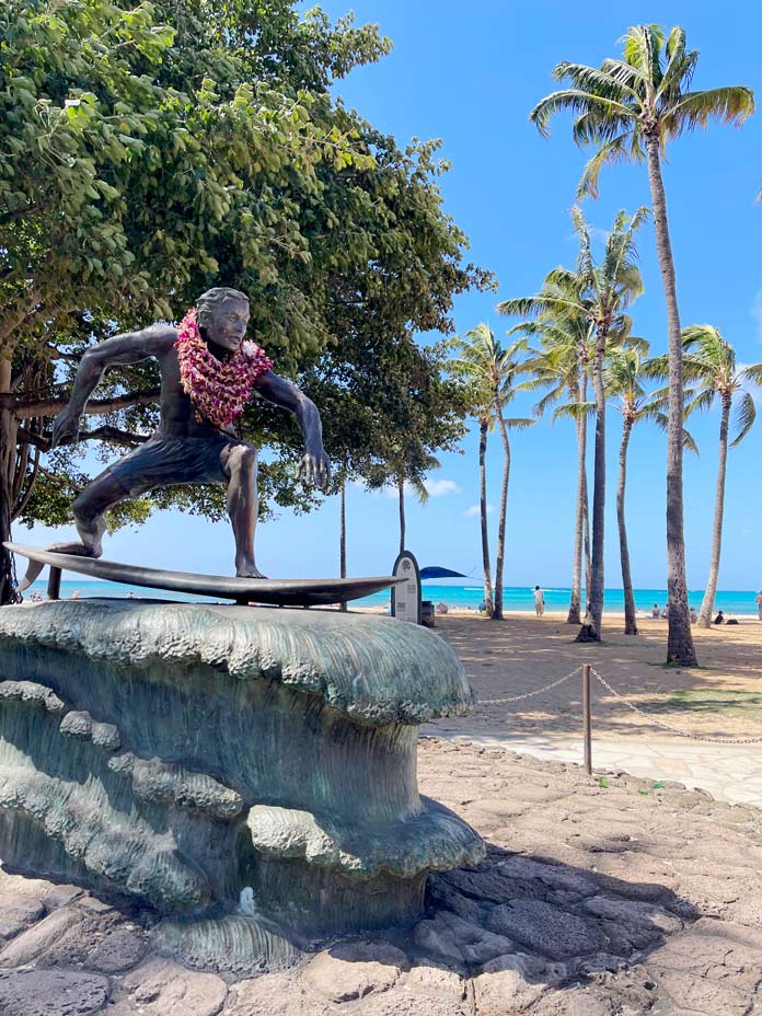 surfende-statue-mit-hawaiikette-am-strand-mit-palmen-auf-oahu