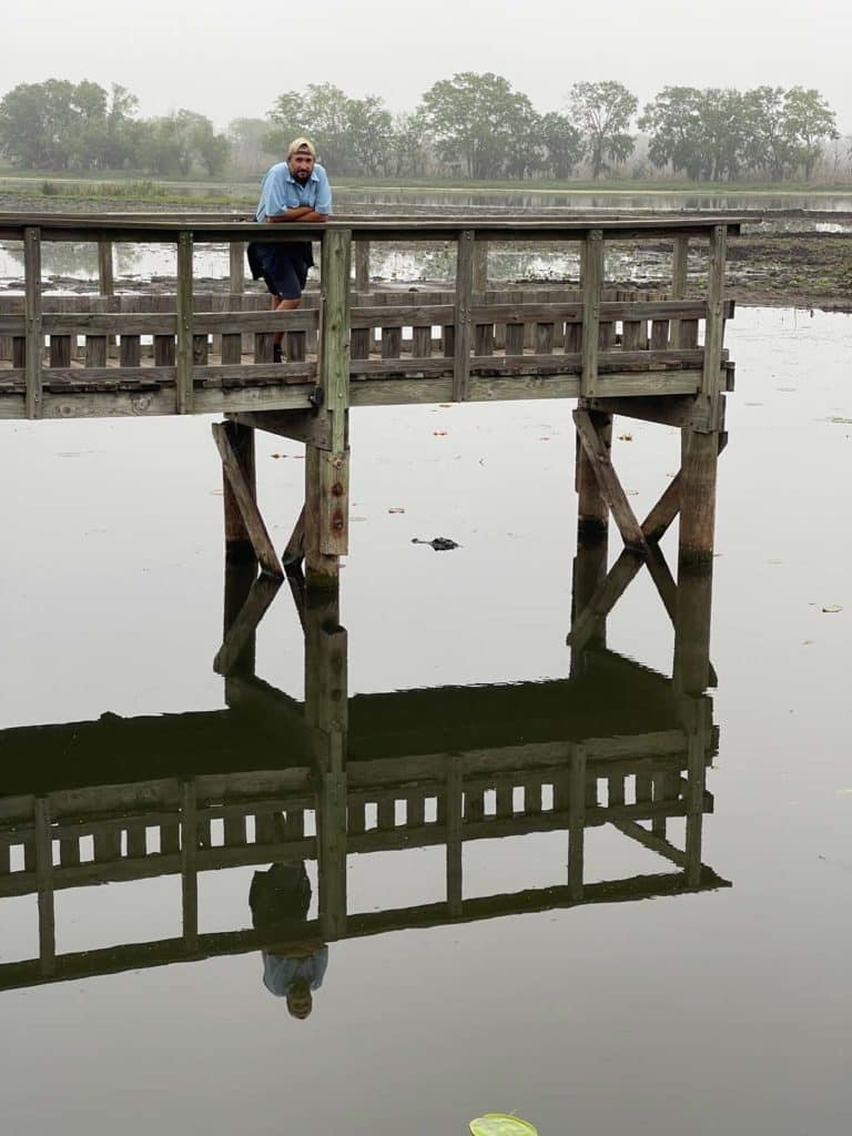 mann-steht-auf-holzsteg-unter-ihm-im-wasser-ein-alligator