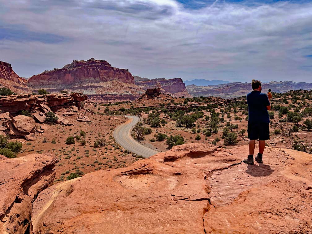 mann-steht-auf-sandsteinfels-und-fotografiert-die-sich-windende-strasse-in-roter-landschaft