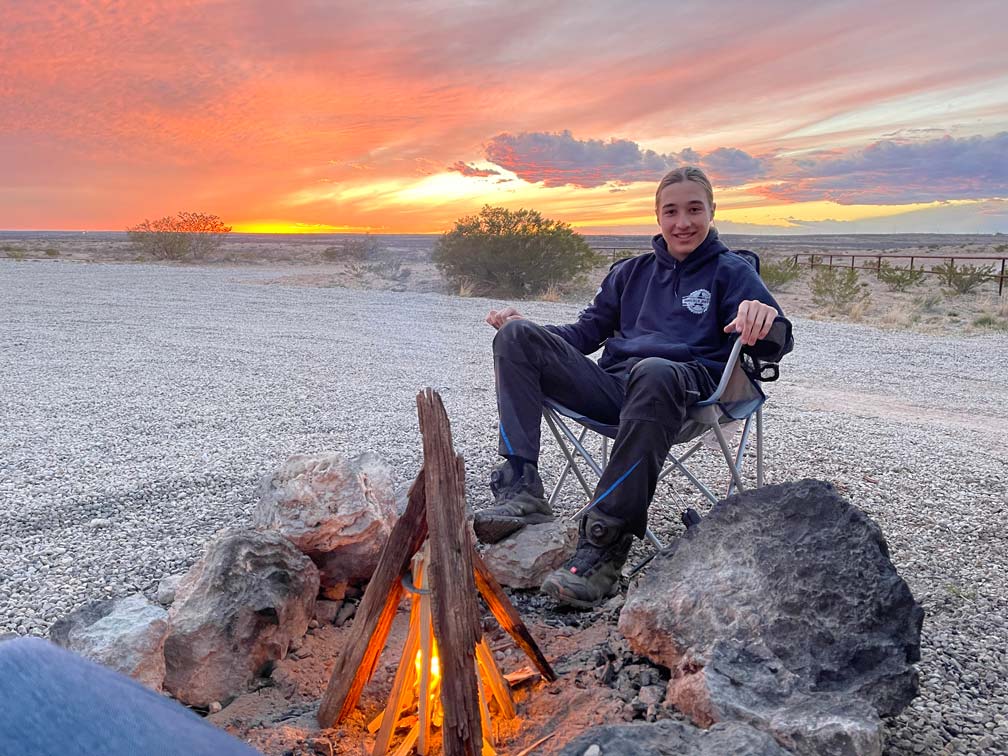 kind-auf-campingstuhl-vor-lagerfeuer-im-hintergrund-leuchtender-sonnenuntergang