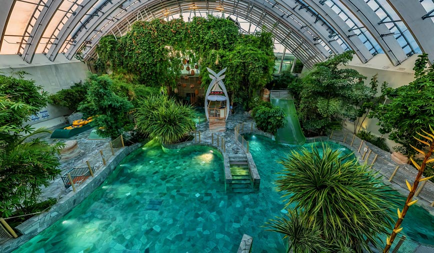 Center Parcs Les Trois Forets - Familienurlaub im Elsass schwimmbad-aqua-mundo-im-center-parcs-les-trois-forets-unter-einer-grossen-kuppel-mit-tropischen-pflanzen-an-der-seite-und-mittig-ein-badehaus-im-bali-stil