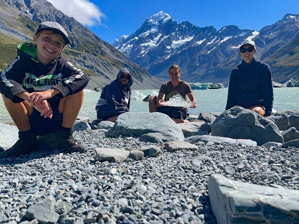 Ăber uns familie-sitzt-am-gletschersee-in-neuseeland-mit-schneebedeckten-bergen-im-hintergrund