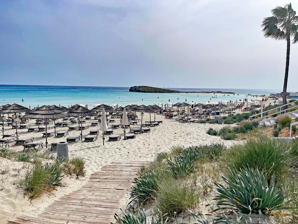 holzsteg-fuehrt-zu-hellem-sandstrand-mit-vielen-liegestuehlen-und-sonnenschirm-am-tuerkisen-meer-mit-blick-auf-kleine-insel-am-nissi-beach-auf-zypern