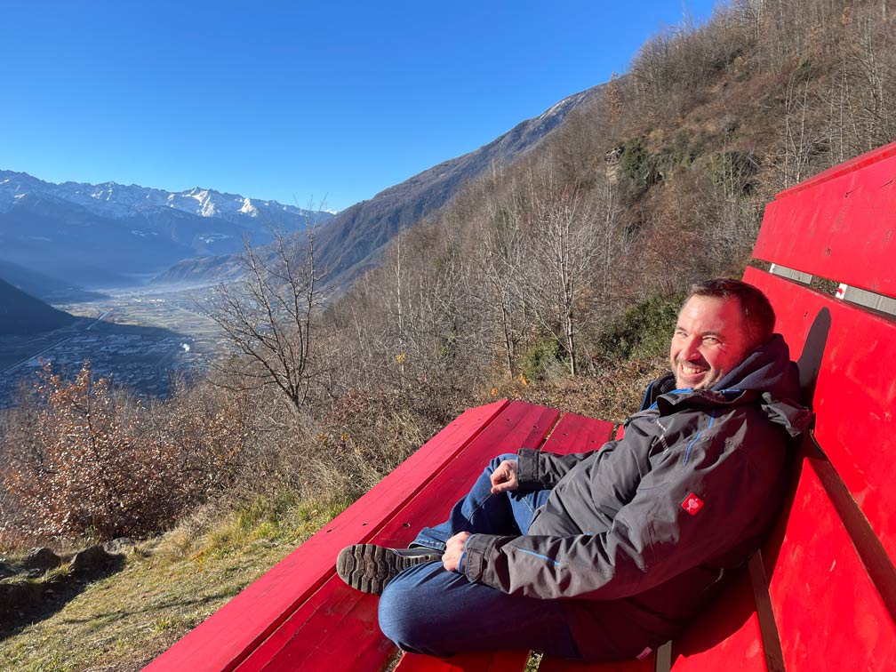 Bernina Express vs. Regionalbahn: Tipps für die perfekte Zugreise durch die Schweizer Alpen Mann-sitzt-auf-großer-roter-bank-mit-blick-auf-berge-und-tirano