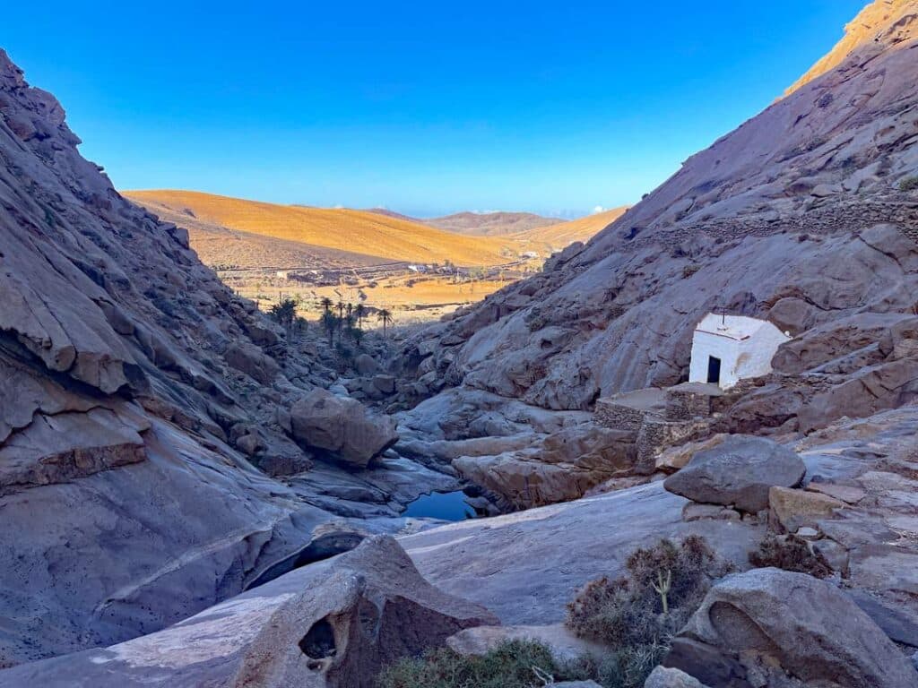 14+ Fuerteventura Sehenswürdigkeiten für den Familienurlaub weisse kirche im barranco de las penitas liegt noch im schatten hinten fast goldschimmernde karge landschaft auf fuerteventura