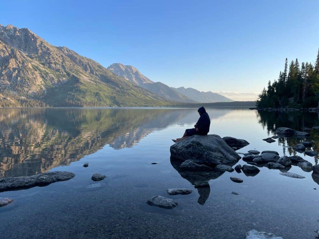 mann sitzt auf stein im jenny lake am fruehen morgen
