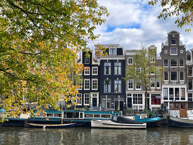 Die Sehenswürdigkeiten in Amsterdam in 3 Tagen herbstlicher baum an gracht in amsterdam mit blick auf schnuecklige häuser und boote davor