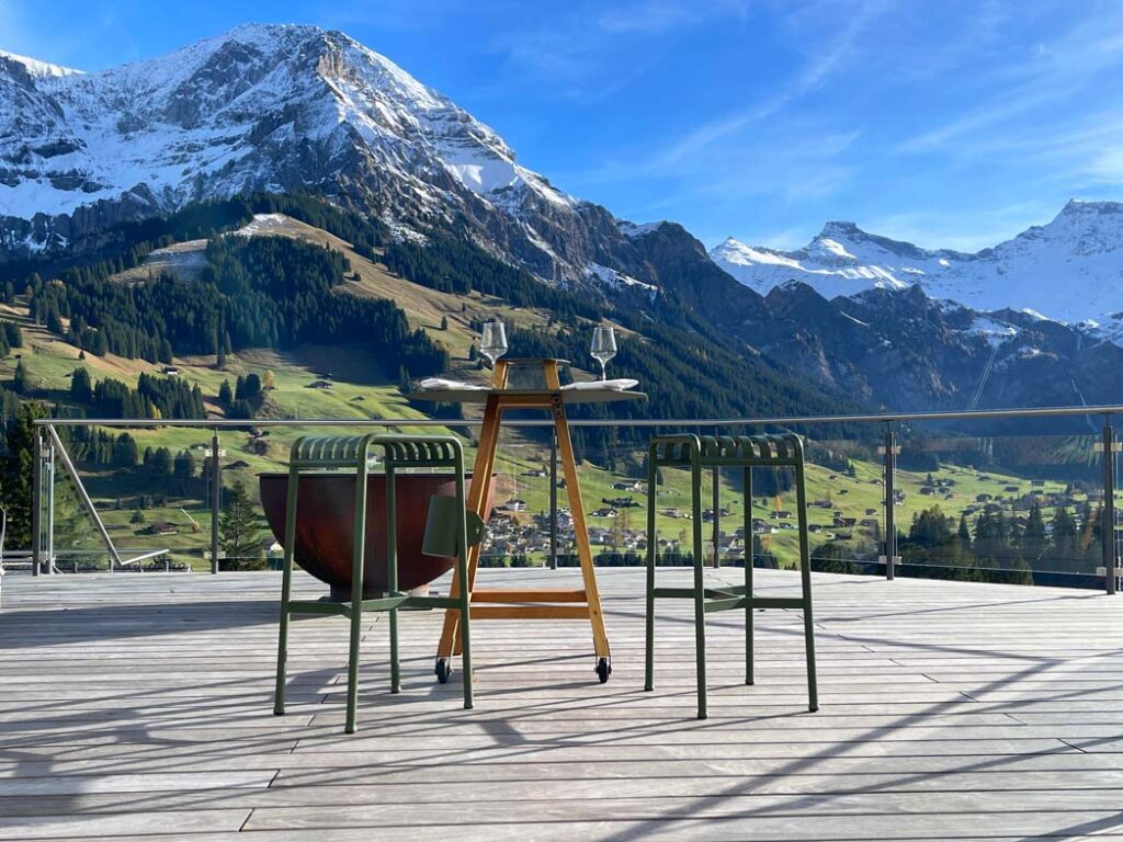 hoher tisch mit hockern und weingläsern neben feuerschale mit blick auf die schweizer berge in adelboden