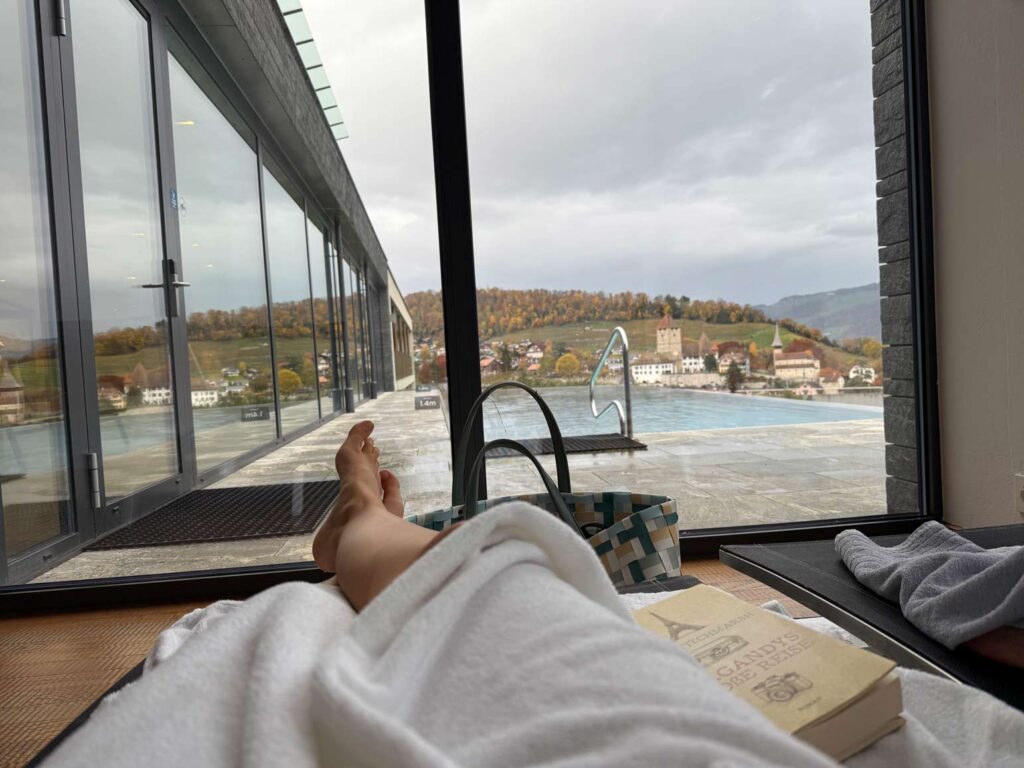 frau im bademantel liegt auf liegestuhl, nur beine zu sehen blick auf inifitypool draußen und spiezer burg