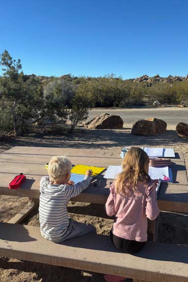 zwei kinder auf sitzbank mit tisch vor ihren arbeitsheften während sie mit ihrer familie auf weltreise sind, sitzend in der natur
