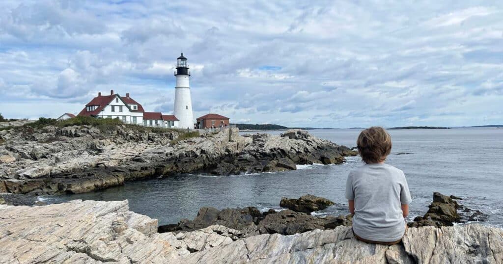 kind sitzt auf felsen und blickt auf leuchtturm in neuengland